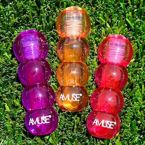 May include: Three colorful, stacked, transparent beauty products in purple, orange, and red. Each stack has four spherical sections and the word "AMUSE" printed on the bottom sphere. The products are on a green, grassy surface.