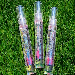 May include: Three clear lip gloss tubes with silver caps are arranged on a bed of green grass. The tubes contain a pink liquid and have the word "PLUMPING" in pink and white text. The brand name "She" is also visible.