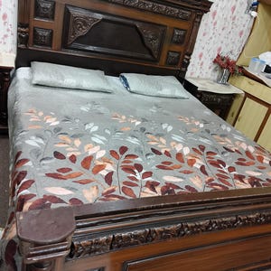 Puede incluir: Un marco de cama de madera con tallas intrincadas y un edredón estampado gris. La cabecera es alta y presenta un diseño decorativo. La cama está hecha con dos almohadas y un edredón gris con un patrón de hojas.