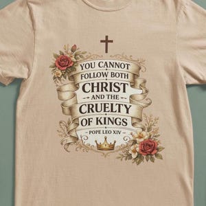 Puede incluir: Camiseta beige con un diseño gráfico que incluye una cruz y la cita "YOU CANNOT FOLLOW BOTH CHRIST AND THE CRUELTY OF KINGS - POPE LEO XIV" en una pancarta decorativa con rosas y una corona.