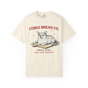 May include: Cream-colored t-shirt featuring a graphic of a corgi dog lying on a wooden cutting board with eggs and wheat. The text "CORGI BREAD CO." is above the dog, and "SMALL LEGS + BIG LOAF ENERGY +" is below.