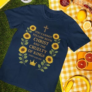 Op de afbeelding: Marineblauw T-shirt met een bloemenkrans van zonnebloemen en bladeren, rondom de tekst "YOU CANNOT FOLLOW BOTH CHRIST AND THE CRUELTY OF KINGS - POPE LEO XIV". Een gouden kroon en kruis zijn ook afgebeeld.