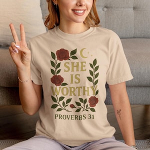 Könnte beinhalten: Beiges T-Shirt mit dem goldenen Schriftzug „SHE IS WORTHY“, umrahmt von einem floralen Design mit roten Rosen und grünen Blättern. Der Text „PROVERBS 31“ steht darunter. Das T-Shirt hat kurze Ärmel.