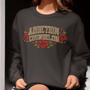 Può includere: Felpa grigio scuro con la scritta "ADDICTION COUNSELOR" in un font leopardato, circondata da rose rosse e foglie verdi. La felpa è a girocollo.