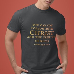 May include: Dark gray t-shirt with gold text. The text reads "YOU CANNOT FOLLOW BOTH CHRIST AND THE CRUELTY OF KINGS -POPE LEO XIV-". The shirt is a crew neck and has short sleeves.