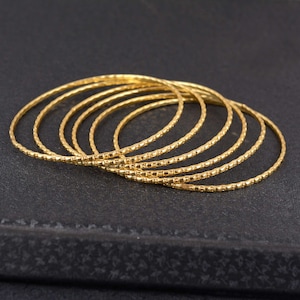 Può includere: Un insieme di sette braccialetti color oro. Ogni braccialetto presenta un motivo strutturato e puntinato. I braccialetti sono impilati, creando un effetto a strati. Lo sfondo è una superficie scura e strutturata.