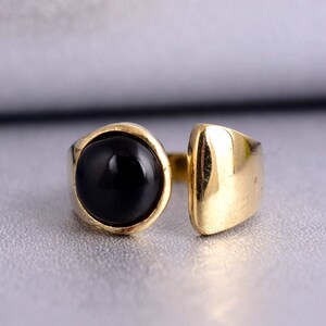 Könnte beinhalten: Ein goldfarbener Ring mit einem großen, runden, schwarzen Stein. Der Ring hat ein offenes Design, wobei der Stein auf einer Seite in einer kreisförmigen Lünette und auf der anderen Seite in einem rechteckigen Band gefasst ist. Der Ring ist vor einem hellgrauen Hintergrund platziert.