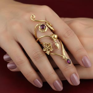 May include: A gold-toned ring with a floral and leaf design, featuring a central flower and two small purple gemstones. The ring wraps around the finger with an elegant, nature-inspired aesthetic.