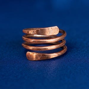 May include: A copper-coloured metal ring featuring a spiral design with three distinct coils. The surface of the metal has a hammered, textured finish, and the ends of the spiral are flattened and rounded, creating a continuous wrap-around band.
