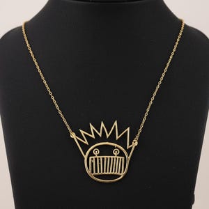 Puede incluir: Collar dorado con un colgante con diseño de corona. El colgante tiene una cara estilizada con una corona encima. El collar se muestra en un maniquí negro.