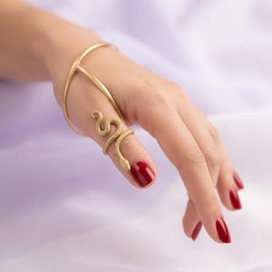 May include: A gold-toned hand bracelet featuring a connected ring design. The piece includes a smooth, thin metal band that wraps around the wrist and back of the hand, leading to a textured snake-shaped ring that coils around the index finger.