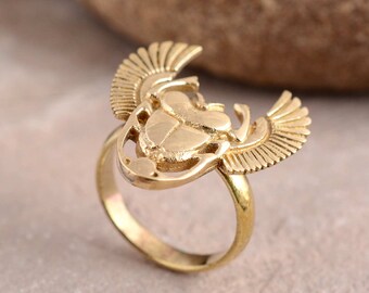 May include: A gold-coloured ring featuring an Egyptian scarab beetle design with outstretched wings. The ring has a detailed, three-dimensional appearance, with the beetle at the centre and wings extending outwards. The ring is set against a neutral background.