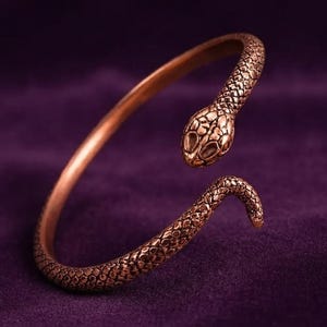 May include: A copper-toned metal cuff bracelet shaped like a coiled snake. The bracelet features a detailed, textured scale pattern along the entire length of the body, with a defined head at one end and a tapered tail at the other end.