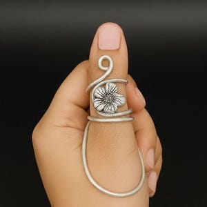 May include: A silver-colored ring featuring a floral design. The ring wraps around the finger with a flower at the center and a decorative swirl at the top. The ring is worn on a finger against a dark background.