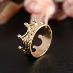 Può includere: Un anello a corona dorata con una fascia strutturata e tre punte rialzate. L'anello è posto su uno sfondo scuro con accenti rosa e verdi sfocati.