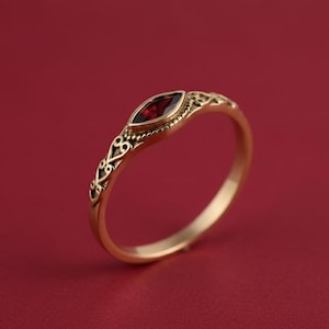 Pode incluir: Um anel dourado com uma pedra preciosa vermelha escura em forma de marquesa. A banda apresenta um design de filigrana ornamentado. O anel está sobre um fundo vermelho liso, destacando os detalhes e a cor da joia.