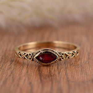 Puede incluir: Un anillo dorado con una piedra preciosa roja oscura de forma ovalada en el centro. La banda presenta intrincados adornos decorativos. El anillo se encuentra sobre una superficie de madera cálida, resaltando su estética vintage.