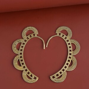 May include: A pair of gold-colored ear cuffs with a unique design. Each cuff features a curved shape with a series of small circles and dots, creating a decorative pattern. The earrings are displayed against a reddish-brown background.