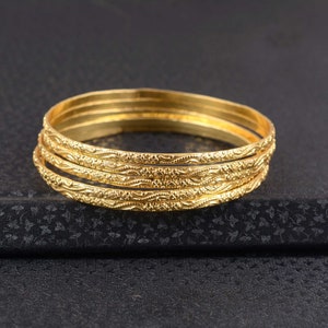 Può includere: Quattro braccialetti dorati con intricati motivi floreali. I braccialetti sono impilati, mostrando il loro design dettagliato e la finitura lucida. I gioielli sono posti su uno sfondo scuro, evidenziando il tono caldo dell'oro.