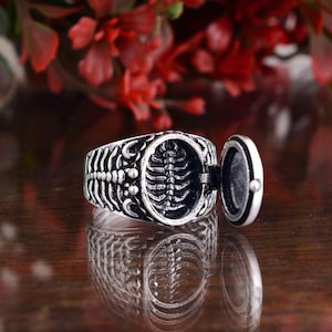 May include: A silver-colored ring with a skeleton design. The ring has an openable compartment revealing a miniature skeleton. The ring is on a wooden surface with red flowers in the background.