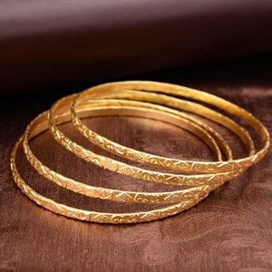 Peut inclure: Quatre bracelets dorés avec des motifs floraux et géométriques complexes. Les bracelets sont empilés sur une surface marron, avec un arrière-plan flou. Les bijoux sont en métal brillant et réfléchissant.