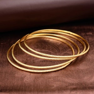 May include: A collection of gold bangles, each with a textured, dotted design. The bangles are stacked, showcasing their circular shape and reflective surface. The background is a dark brown, creating a contrast with the gold.