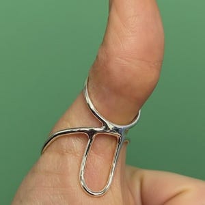Könnte beinhalten: Ein silberfarbener Ring mit einem einzigartigen, offenen Design. Der Ring hat ein gebogenes Band, das den Finger umschließt, mit einem länglichen, U-förmigen Element. Der Ring wird auf einem Daumen vor grünem Hintergrund gezeigt.