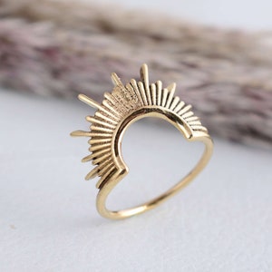 Puede incluir: Un anillo dorado con diseño de rayos de sol. El anillo presenta una forma de semicírculo con líneas radiantes, creando una apariencia de sol. El anillo está hecho de un material metálico brillante.