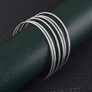 Puede incluir: Una pila de seis pulseras finas de plata. Las pulseras circulares y brillantes descansan sobre una superficie texturizada de color verde oscuro. El fondo es gris oscuro.