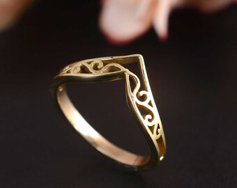 May include: A gold-colored ring with a V-shaped design. The ring features intricate scrollwork along the V-shape and band. The ring is set against a dark background, highlighting its details. This piece is a jewelry item.