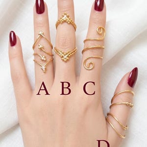 May include: Four gold-toned rings labeled A, B, C, and D. Ring A is a chevron design with small accents. Ring B features two chevron bands with beaded details. Ring C is a spiral wrap design. Ring D is a multi-coil band. All rings are gold-colored.