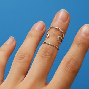 May include: A silver-colored finger ring with a unique design. The ring features two bands connected by curved accents, creating an open, artistic look. The ring is worn on a finger against a solid blue background.