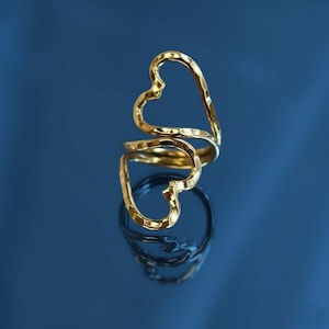 May include: A gold-colored ring with a heart-shaped design. The ring features two open hearts connected by a band, creating a unique and stylish look. The ring is displayed on a reflective surface against a blue background.