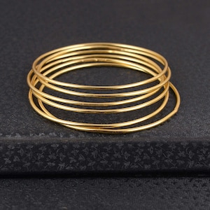 Puede incluir: Una pila de seis pulseras finas de color dorado. Las pulseras son circulares y parecen ser de metal. Están dispuestas de forma ligeramente superpuesta, descansando sobre una superficie oscura.