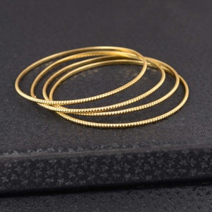 Peut inclure: Ensemble de quatre bracelets dorés. Chaque bracelet présente un design fin et délicat avec un motif texturé et pointillé. Les bracelets sont empilés sur une surface sombre et texturée, créant une esthétique simple et élégante.