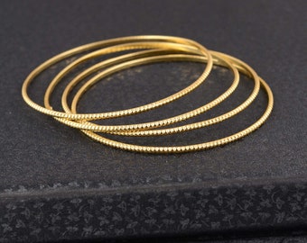 May include: A collection of four gold-colored bangles. Each bangle has a thin, delicate design with a textured, dotted pattern. The bangles are stacked on a dark, textured surface, creating a simple yet elegant aesthetic.