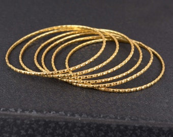 May include: A collection of seven gold-colored bangles with a textured, dotted pattern. The bangles are arranged in a slightly overlapping arc, set against a dark background. The jewelry is simple and elegant.