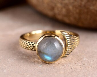 Può includere: Un anello color oro con una gemma rotonda blu chiaro. L'anello ha una fascia strutturata e un design decorativo a forma di ala. La gemma ha un diametro di circa 1,3 cm.
