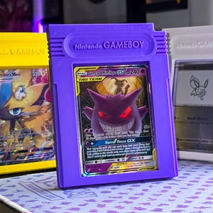 Puede incluir: Tres soportes para cartas con forma de Gameboy en amarillo, morado y blanco, cada uno con una carta Pokémon. El soporte morado muestra una carta Gengar & Mimikyu GX. El texto "Nintendo GAMEBOY" es visible en el soporte morado.
