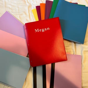 May include: A collection of colorful notebooks and folders, including red, blue, pink, purple, orange, and yellow. The red folder has the name "Megan" printed on it in white. The notebooks have spiral binding.