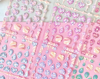 May include: Sheets of decorative stickers with gem shapes, including hearts, teardrops, and stars, in pink, purple, and blue. The stickers have a glittery, iridescent finish and are arranged on a pink background.