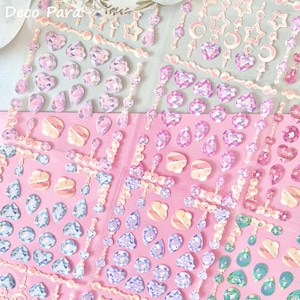 May include: Sheets of decorative stickers featuring various gem shapes, including hearts, teardrops, and stars, in shades of pink, purple, and blue. The stickers have a glittery, iridescent finish and are arranged on a pink background.