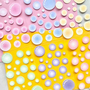 May include: A collection of colorful, bubble-shaped stickers in various sizes. The stickers feature pastel colors like pink, blue, yellow, green, and purple, arranged on pink and yellow backgrounds. These stickers are ideal for scrapbooking and crafts.