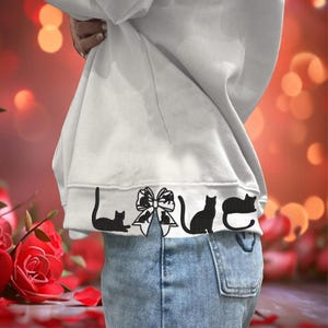 May include: White sweatshirt with a black design of cats forming the word "LOVE" with a bow in the middle. The sweatshirt is being worn by a person and the bottom of the sweatshirt is tucked into blue jeans.