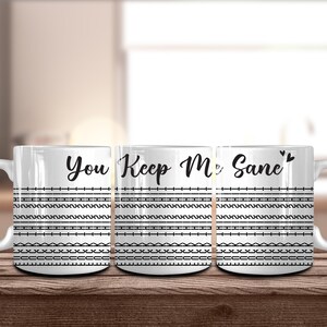 May include: White ceramic mugs with the phrase "You Keep Me Sane" in black script. The mugs feature black horizontal lines and geometric patterns. The mugs are on a wooden surface.