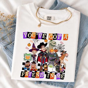 May include: A white t-shirt featuring a graphic print of various animated characters dressed in Halloween costumes. The text "YOU'VE GOT A FRIEND IN ME" is printed in colorful, blocky letters above and below the characters, surrounded by spiderwebs.