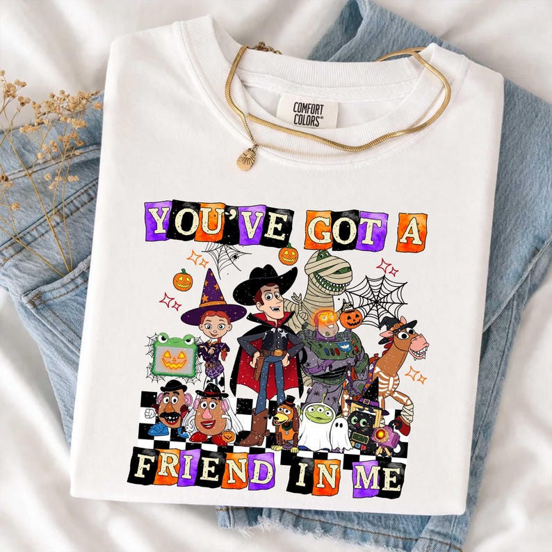 May include: A white t-shirt featuring a graphic print of various animated characters dressed in Halloween costumes. The text "YOU'VE GOT A FRIEND IN ME" is printed in colorful, blocky letters above and below the characters, surrounded by spiderwebs.