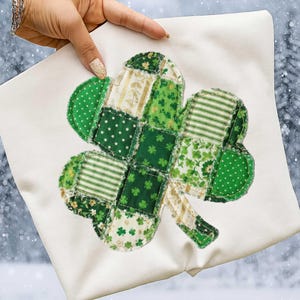 May include: A white square pillow with a large, patchwork shamrock design. The shamrock is composed of various green fabrics, including polka dots, stripes, and floral patterns. The pillow is held up by a hand with a gold nail polish.