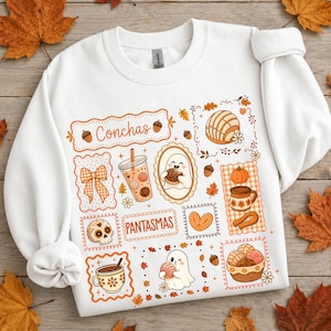 May include: A white long-sleeve sweatshirt featuring a graphic print with the text Conchas and Pantasmas. The design includes illustrations of ghosts, concha bread, coffee cups, pumpkins, acorns, and bows in shades of orange and brown.