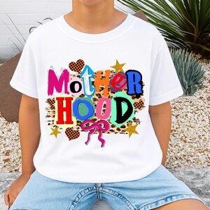 May include: White t-shirt with the word "MOTHERHOOD" in colorful, playful letters. The design includes hearts, stars, and a bow, all set against a leopard print background. A fun and vibrant graphic tee.